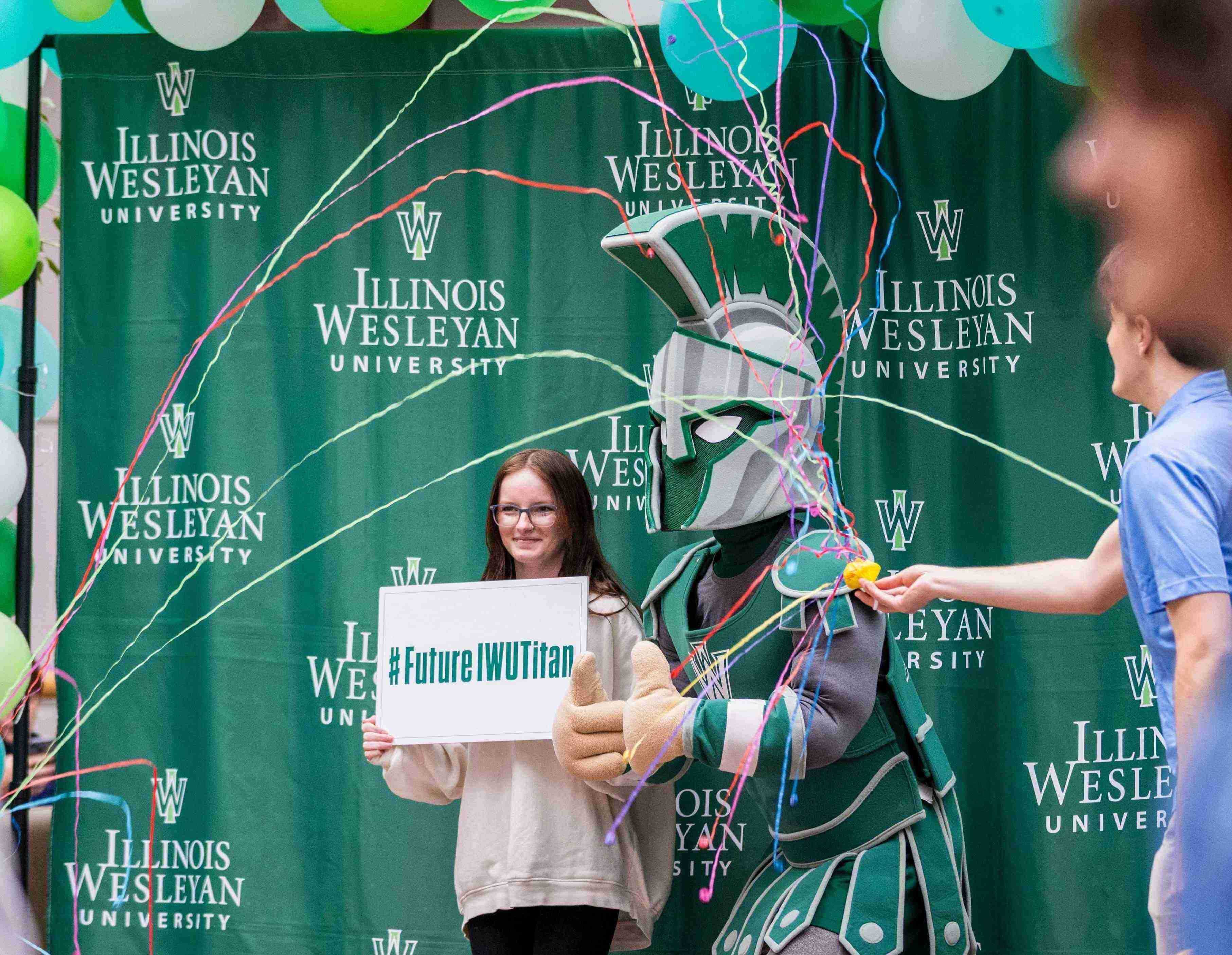 Student holding a #FutureTitan sign in front of a green backdrop with Tommy the Titan as confetti flies by