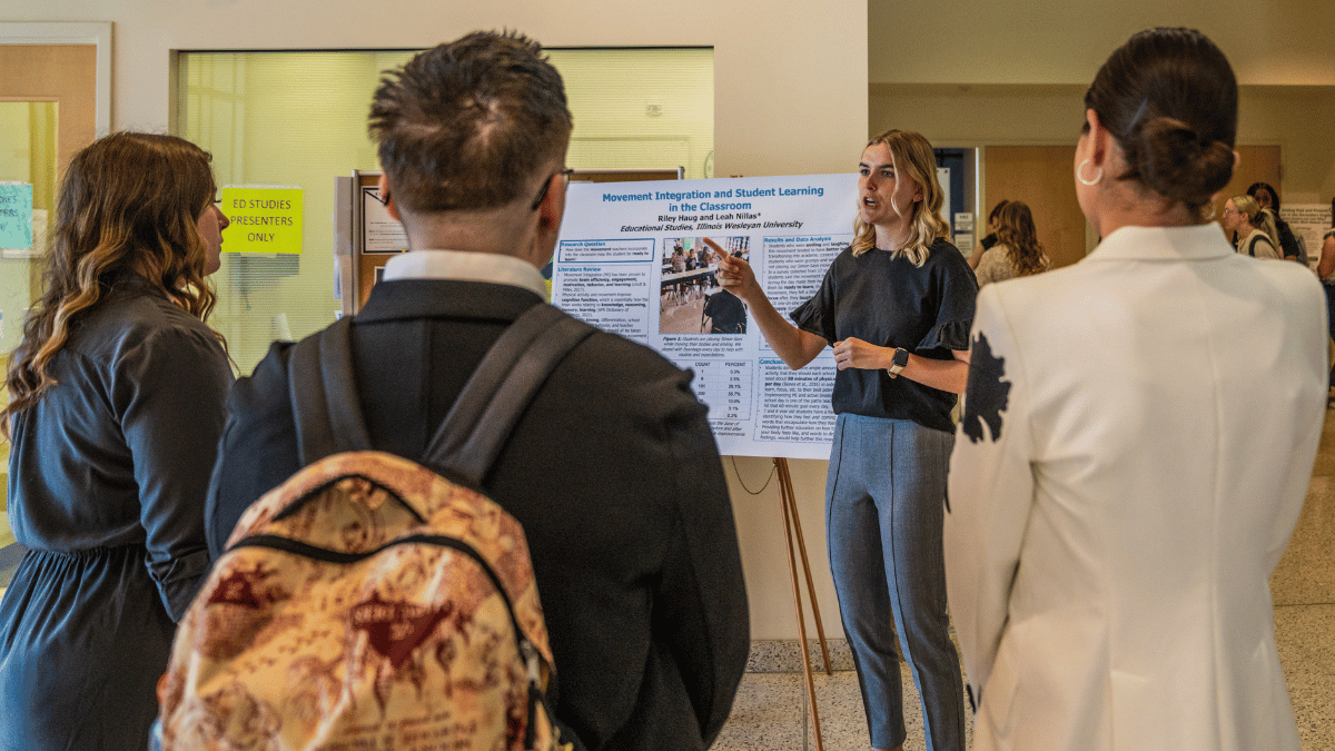 JWP Conference Three people stand listening to a student presenting research on a poster in IWU's State Farm Hall