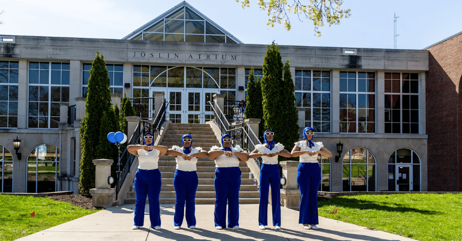 Five IWU students stand in front of Joslin Atrium with elbows raised in a dance, wearing blue and white as part of the Zeta Phi Beta sorority chartering ceremony